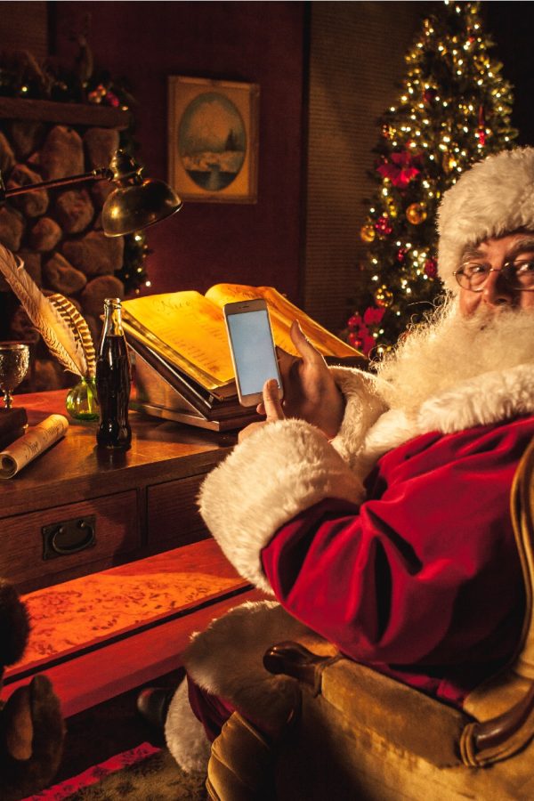 Santa holds a smartphone at his desk with Christmas decor in the background