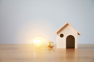 lit lightbulb is on its side next to a wooden model of a house.